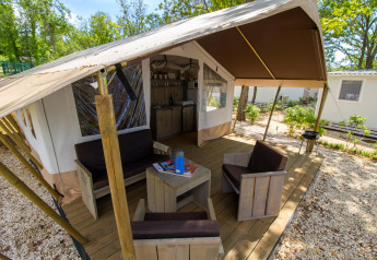 Tenda safari con veranda in legno, area relax e cucina, immersa nella natura e circondata da alberi.