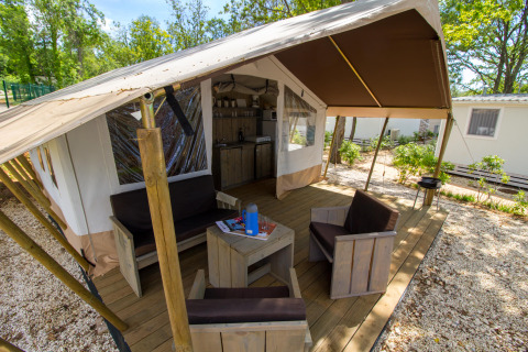Tenda safari con veranda in legno, area relax e cucina, immersa nella natura e circondata da alberi.