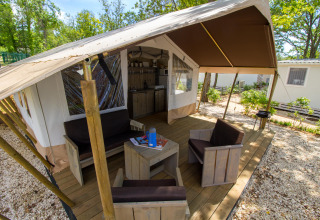 Safari-Zelt mit Holzterrasse, gemütlichen Sitzmöbeln und Küche, umgeben von Bäumen und anderen Zelten.