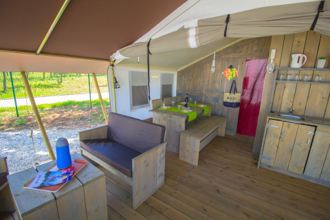 Interior of a safari tent featuring wooden furniture, a dining area, sofa, and small kitchenette in nature.