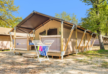 Safari telt Lodge med aircondition, placeret blandt træer, med terrasse og tørrestativ foran under blå himmel.