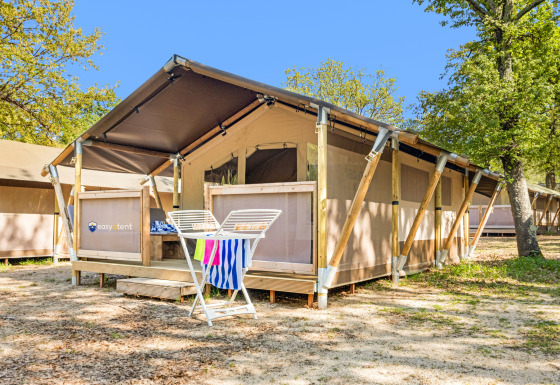 Safari telt Lodge med aircondition, placeret blandt træer, med terrasse og tørrestativ foran under blå himmel.