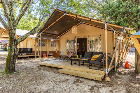 Lodge de tienda safari con terraza de madera, sillones cómodos y sofá, rodeada de árboles y naturaleza.