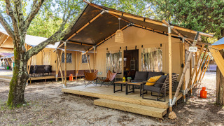 Lodge de tienda safari con terraza de madera, sillones cómodos y sofá, rodeada de árboles y naturaleza.