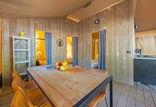 Binnenaanzicht van een safaritent met houten wanden, eettafel, stoelen en zichtbare slaapkamers.