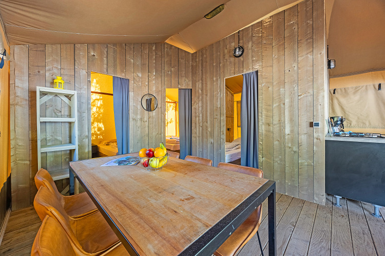 Interior view of a safari tent featuring wood-paneled walls, dining table, chairs, and visible bedrooms.