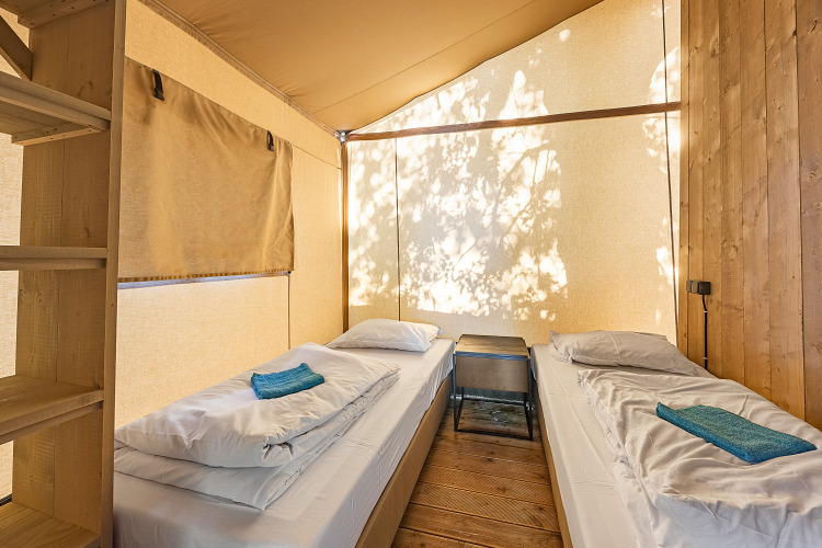Interior of a safari tent with two single beds, white bedding, wood floor, and natural light filtering in.