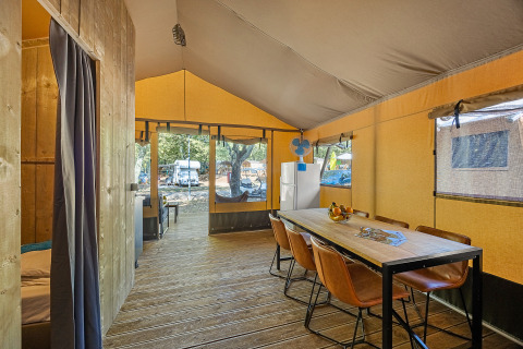 Interior view of a spacious safari tent with wooden floor, dining table, chairs and a refrigerator inside.