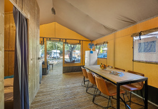 Interior view of a spacious safari tent with wooden floor, dining table, chairs and a refrigerator inside.