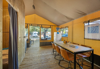 Interior view of a spacious safari tent with wooden floor, dining table, chairs and a refrigerator inside.