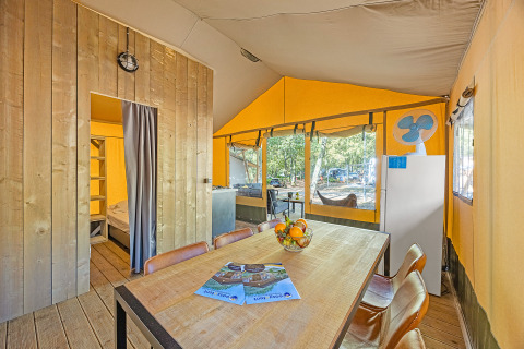 Interior of a safari tent with a dining table, fruit bowl, fridge, and view outside to a wooded campsite.