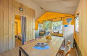 Interieur van een safaritent met eettafel, fruitschaal, koelkast en uitzicht naar buiten op de camping.