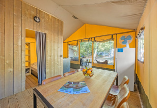 Binnenkant van een safaritent met eettafel, fruitschaal, koelkast en zicht op het bos buiten.