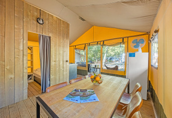 Interior of a safari tent with a dining table, fruit bowl, fridge, and view outside to a wooded campsite.