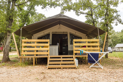 Luxuriöses Safarizelt mit überdachter Terrasse und Holzbalkon, eingebettet in eine grüne Umgebung.
