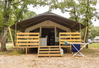 Luxury safari tent with covered porch, wooden railings and outdoor seating, set among green trees.