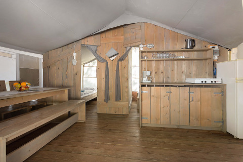 Interno di una tenda safari di lusso con arredi in legno, tavolo da pranzo, angolo cottura e camera.