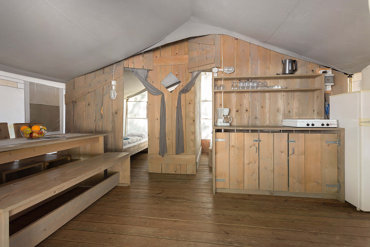 Interior of a luxury safari tent featuring wooden furnishings, dining table, kitchenette, and bedroom access.