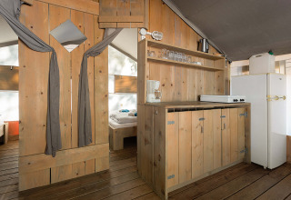 Vista interna di una tenda safari di lusso con pavimento in legno, angolo cucina e zona letto visibile.