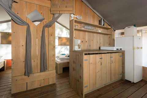 Vue intérieure d'une tente safari de luxe avec plancher en bois, coin cuisine et espace chambre visible.