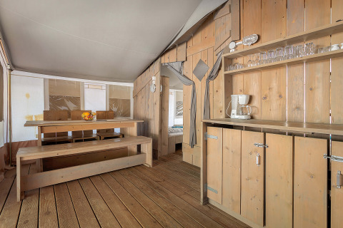 Interno della Safari Tent Luxury con panche e tavolo in legno, cucina attrezzata e stoviglie per comfort.