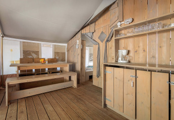 Interior of Safari Tent Luxury featuring wooden benches, dining table, kitchenette and glassware for comfort.