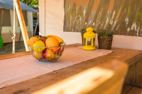 Cesto di frutta su tavolo in legno in una tenda safari di lusso, con lanterna gialla e piante in vaso.
