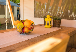 Cesto di frutta su tavolo in legno in una tenda safari di lusso, con lanterna gialla e piante in vaso.