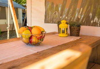 Cesto di frutta su tavolo in legno in una tenda safari di lusso, con lanterna gialla e piante in vaso.
