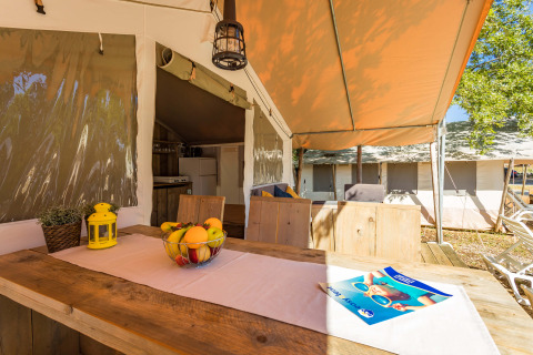 Zona pranzo all'aperto in tenda safari di lusso con mobili in legno, ciotola di frutta e atmosfera accogliente.