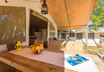 Zona pranzo all'aperto in tenda safari di lusso con mobili in legno, ciotola di frutta e atmosfera accogliente.