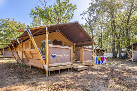 Tente safari sur un camping, avec terrasse en bois et étendoir à serviettes, entourée de nature.
