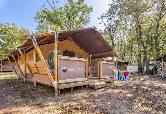Tente safari sur un camping, avec terrasse en bois et étendoir à serviettes, entourée de nature.