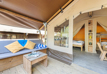 Safari-Zelt mit Holzsofa, Kissen, kleiner Tisch, Essbereich und sichtbaren Betten im Hintergrund.
