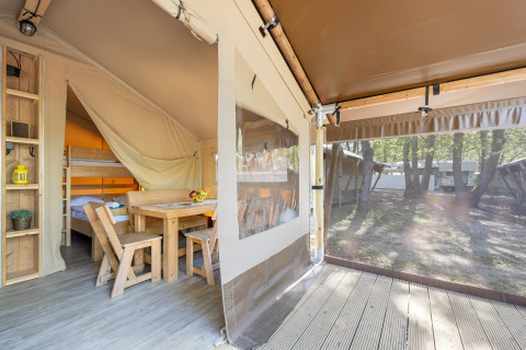 Interior de tienda safari con muebles de madera, litera y ventana transparente con vista al bosque exterior.