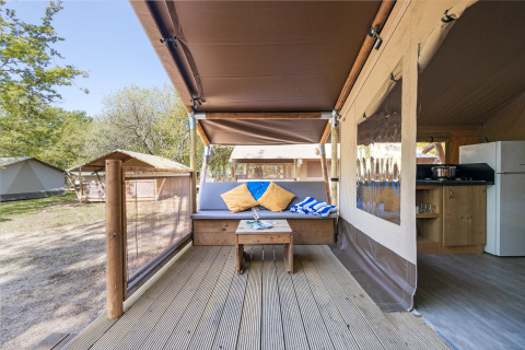 Outdoor seating area of a safari tent with a cushioned bench, table, and kitchen in a natural setting.