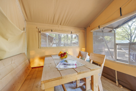 Intérieur d'une tente safari avec coin repas, table en bois, bancs et fenêtres donnant sur la nature.