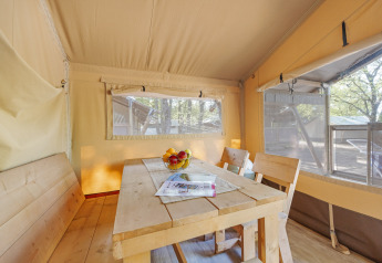 Interior of a safari tent dining area with wooden table, benches, and large windows showing the outdoors.