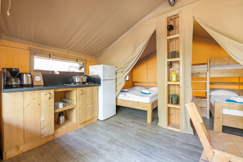 Interno di una tenda safari con angolo cottura, frigorifero, letto matrimoniale e letti a castello in legno.