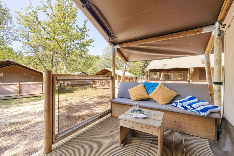 View from a safari tent porch with a cushioned bench, table, pillows, and striped towels amid trees.