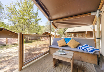 View from a safari tent porch with a cushioned bench, table, pillows, and striped towels amid trees.