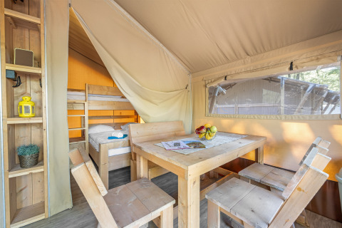 Innenansicht eines Safarizelts mit Holztisch, vier Stühlen, Regalen und einem Etagenbett im Hintergrund.