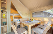 Interieur van een safaritent met houten eettafel, stoelen, open kast en zichtbaar stapelbed.