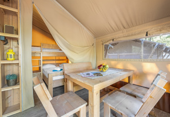 Interno di una tenda safari con tavolo da pranzo in legno, sedie, scaffali e letto a castello.