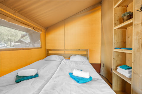 Intérieur d'une tente safari avec deux lits simples, étagères en bois et serviettes soigneusement pliées.