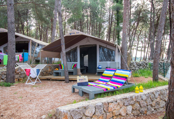 Luxury safari tent in a wooded area with colorful lounge chairs, a deck, and drying laundry outside.