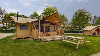 Glampingverblijf met luxe tent, houten terras en picknickbank op een groen grasveld.