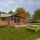 Et glamping-sted med et luksuriøst telt, terrasse og bænk omgivet af grønne træer under en blå himmel.