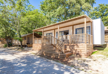 Mobile home Pula met houten veranda, gelegen in een groene omgeving en omgeven door bomen en natuur.