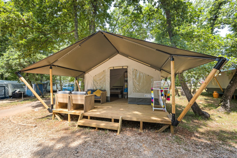 Tienda safari con terraza de madera y asientos exteriores, ubicada en un bosque rodeado de árboles verdes.